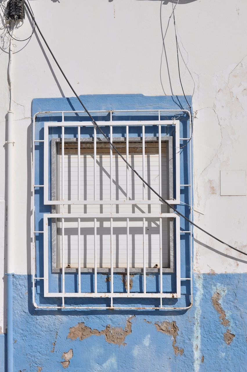 Au Portugal, une fenêtre fermée avec une grille, le mur est bleu et blanc mais très vétuste et des câbles électriques sont apparents.